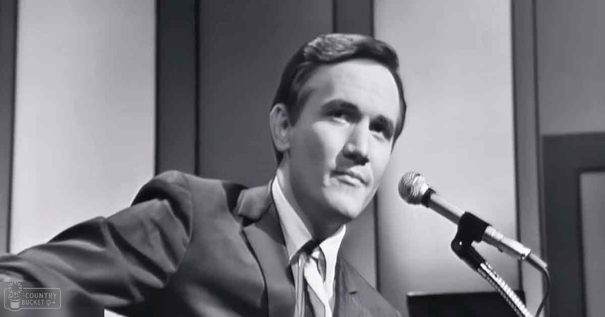 A middle-aged adult male in formal attire appears to be giving a speech or presentation in an indoor setting with paneling and a doorframe, suggesting it might be from the mid-20th century.