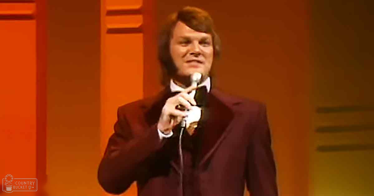 An adult male performer in a dark suit with a white shirt, holding a microphone on an indoor stage with orange paneling and warm lighting.