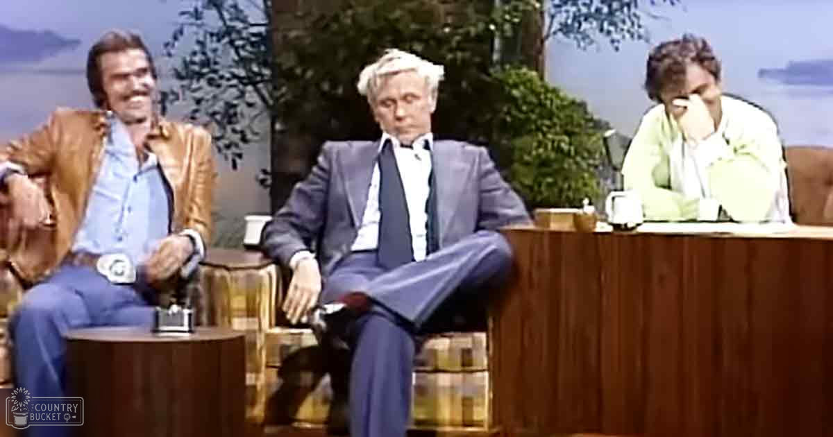 A daytime talk show set mimicking an outdoor scene with three individuals seated against a backdrop of trees and water.
