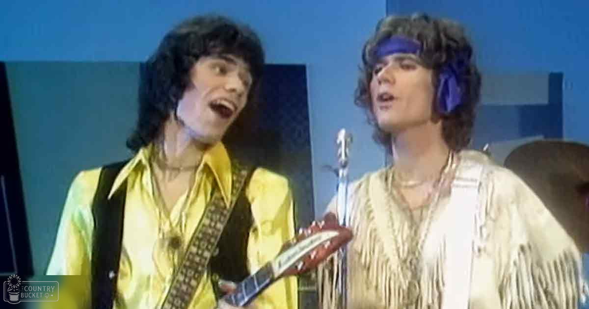 A musical performance scene with two young adult male musicians, one wearing a yellow satin shirt and the other a white fringe jacket, performing on stage under blue lighting.