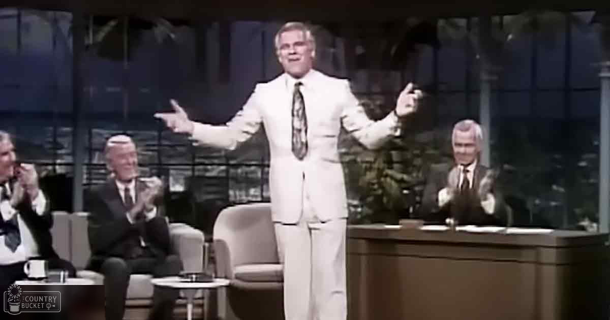 A talk show setting with multiple adult males seated and clapping, dressed in formal suits against a backdrop of large windows showing a cityscape at night or dusk.