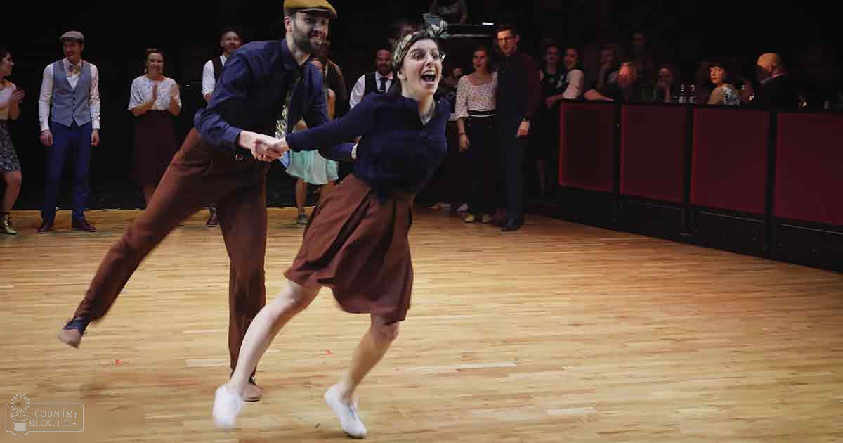 A young adult couple dancing in a dance hall or studio with wooden flooring and an audience watching dressed in early 20th-century attire. The scene suggests a historical reenactment or themed party, illuminated by even lighting.