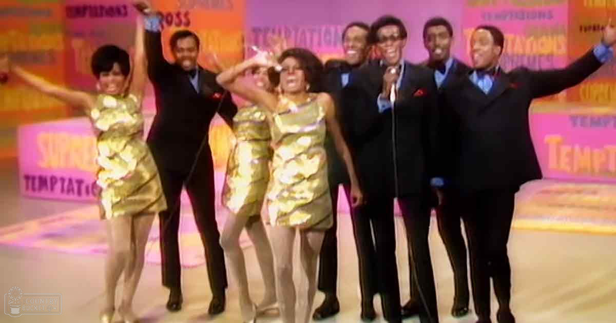 A performance group from the 1960s, dressed in metallic gold dresses and black suits with patterned ties, stands on a stage or studio set with colorful 'Temptations' banners behind them.