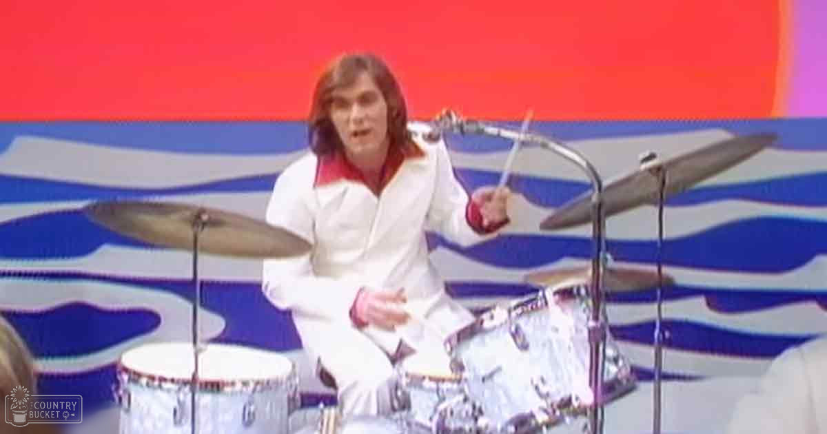 A young adult male drummer wearing a white and red zip-up jacket performs on stage with a drum set, under bright indoor lighting.