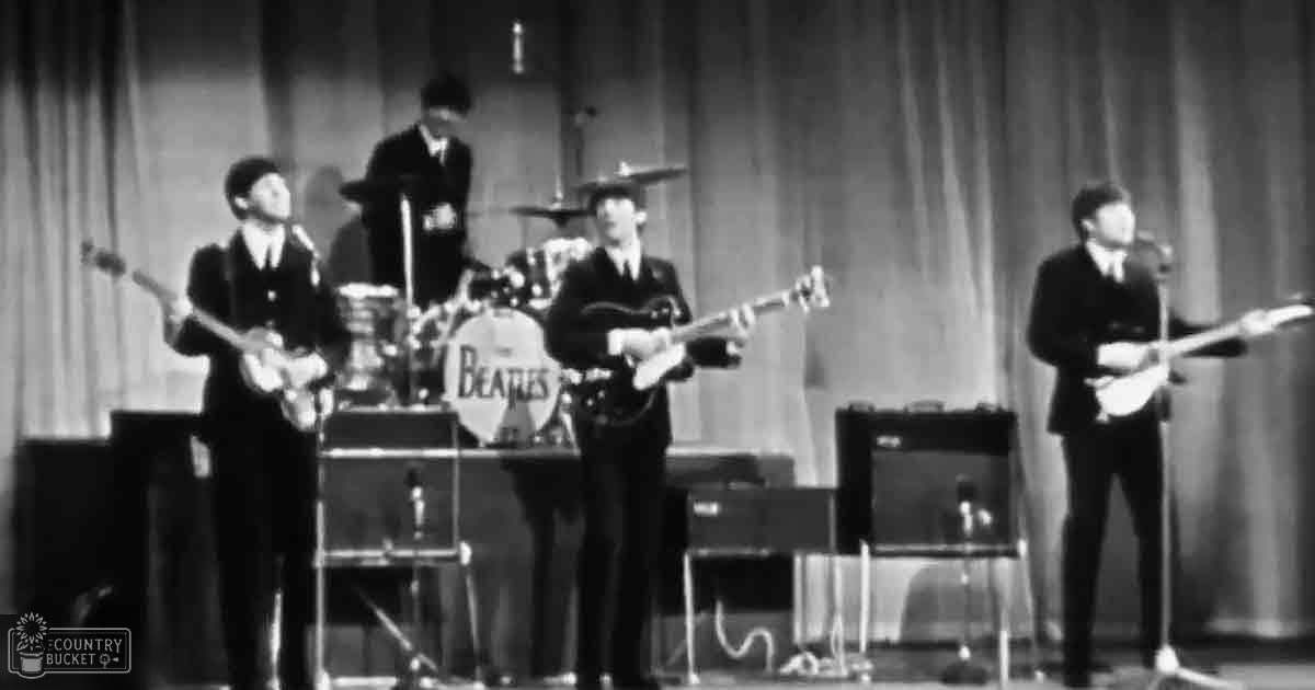 A musical performance by a band from the 1960s with young adult males dressed in dark suits and white shirts, with a drum set prominently featured on stage under bright lighting against a plain curtain backdrop.