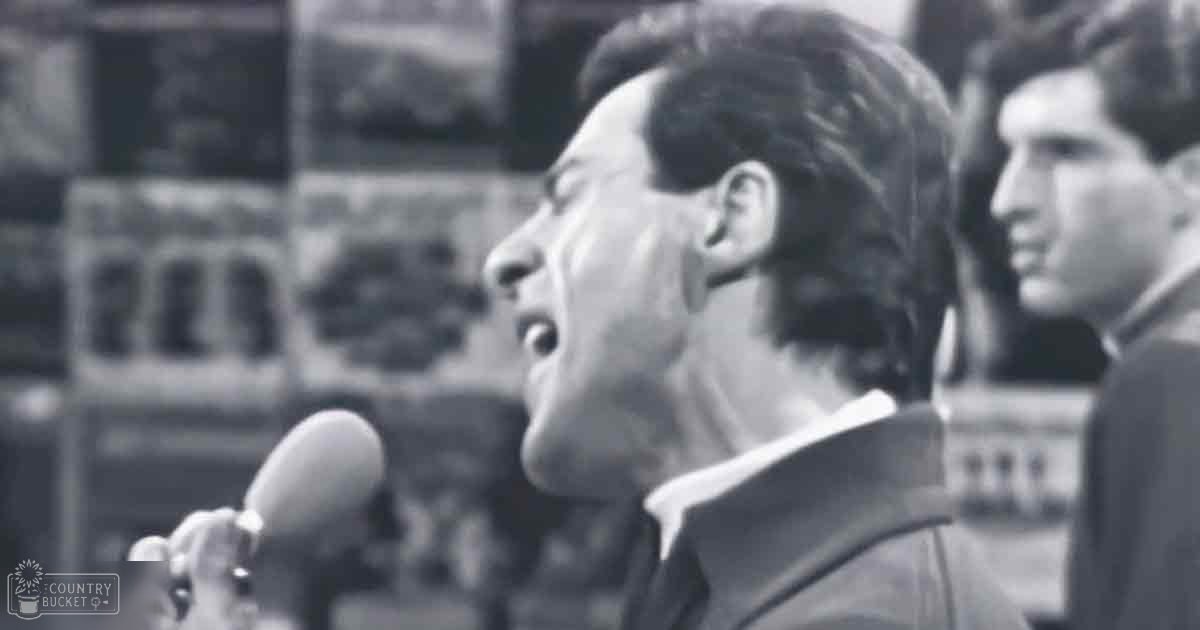 An adult male in a dark suit jacket speaks into a microphone outdoors on what appears to be late afternoon with natural light, possibly at a government or public building.