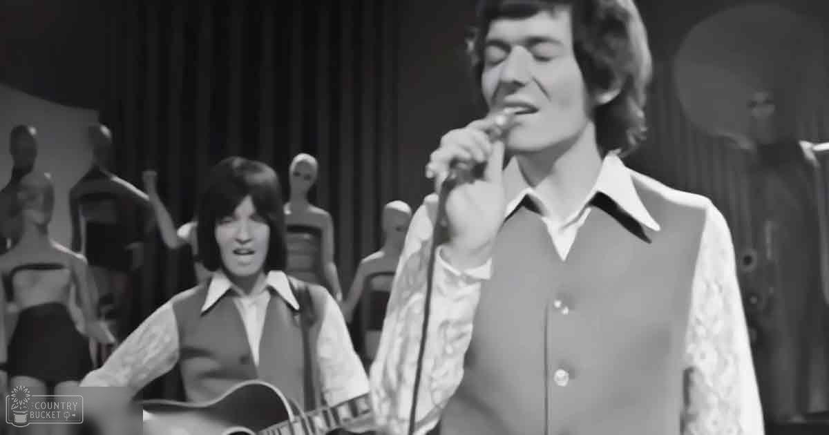 A young adult male singer wearing a white lace-patterned shirt with dark vest stands on stage holding a microphone, surrounded by mannequins and curtains in black-and-white lighting.