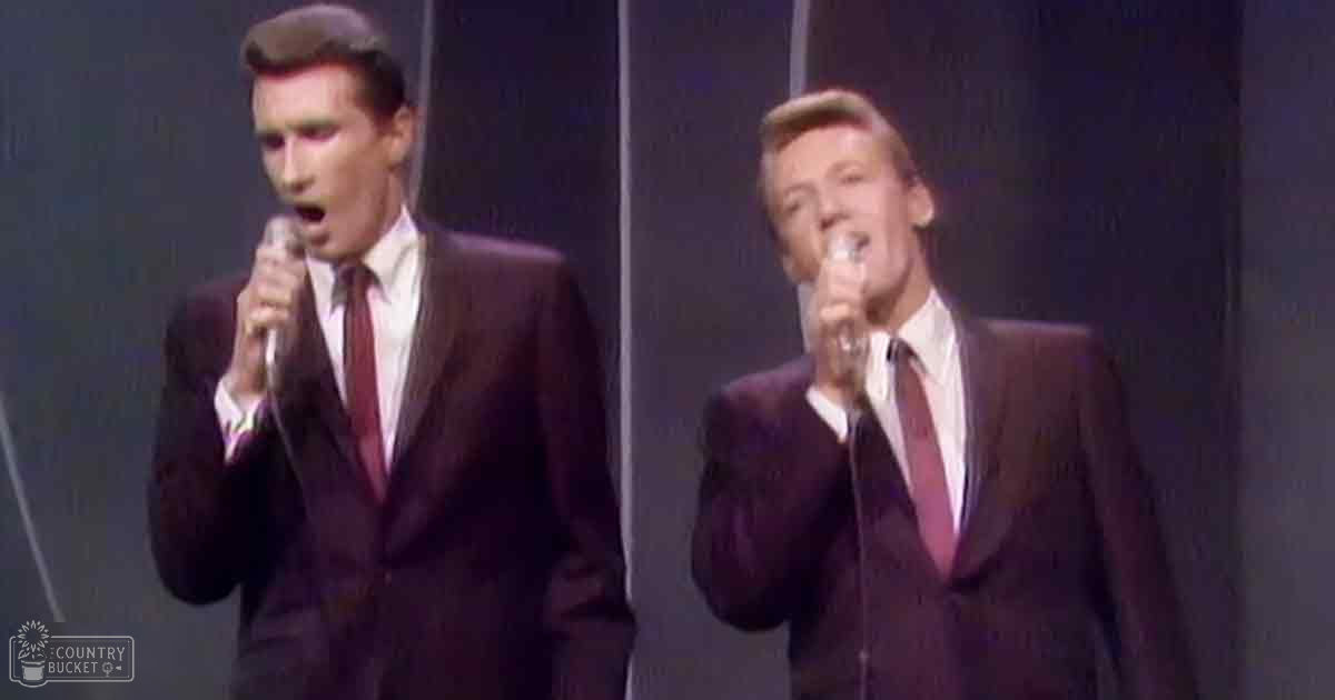 A musical performance scene with two young adult male singers on stage, wearing dark suits and white dress shirts with red ties, set against a backdrop of dark curtains and lighting equipment.