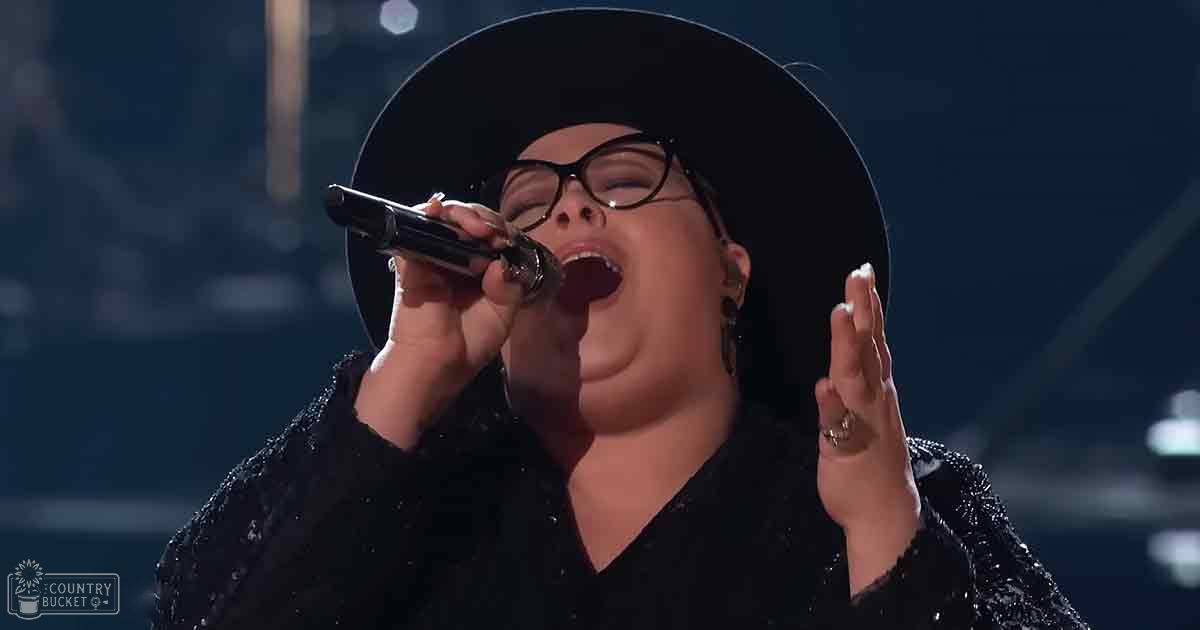 A female adult singer wearing a black, intricately patterned V-neck garment with long fitted sleeves, performing on stage under spotlights.