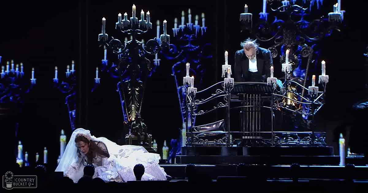 A theatrical scene with a grand hall or castle interior, with an adult female in a white wedding gown holding flowers and an older male in a dark suit wearing glasses, surrounded by ornate furniture including a grand piano and chandeliers.