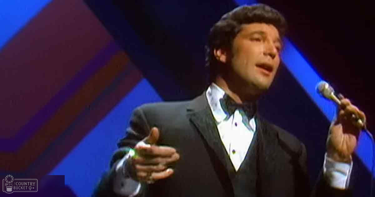 Adult male performer wearing a dark tuxedo with bow tie, holding a microphone against stage lighting with blue and red hues.