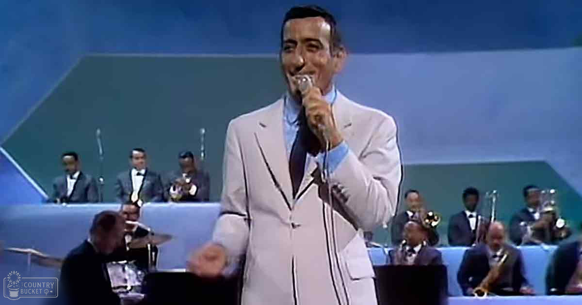 A man singing in a white suit with an orchestra on stage under bright lighting, likely from the mid-20th century.