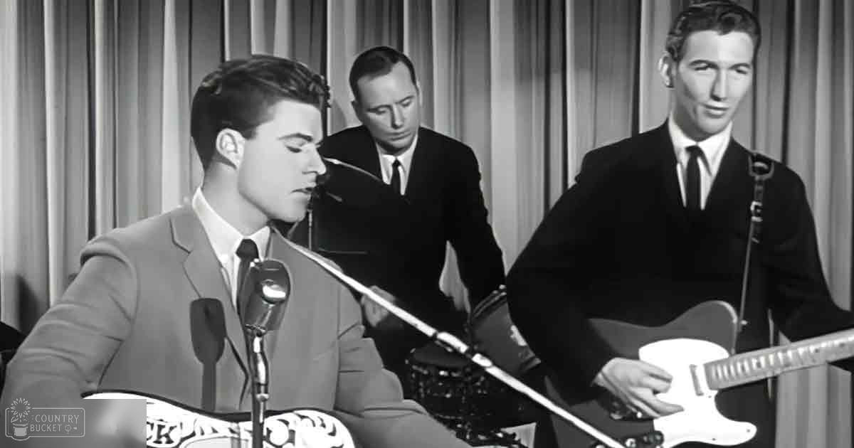 A young adult male singer in a light-colored suit jacket, white shirt with dark tie, and wristwatch performs into a microphone while another adult male in a dark suit stands behind him. A third adult male also in a dark suit holds a guitar labeled 'RICK' in front of the drum set.