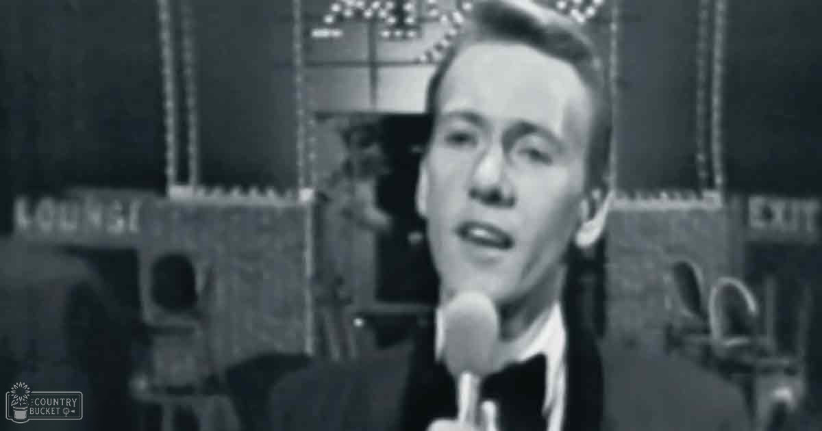 An adult male singer wearing a dark suit jacket with a white shirt and dark tie, performing into a microphone in an indoor lounge setting.