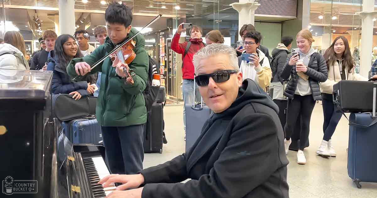 A middle-aged man in a black hoodie plays piano alongside a teen boy playing violin amidst travelers in an airport terminal, winter or late fall setting with muted tones and artificial lighting.
