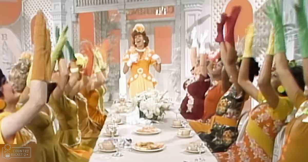 A theatrical or musical performance setting with multiple individuals in yellow dresses, gloves, and a long table set for food, against decorative stage backdrops.
