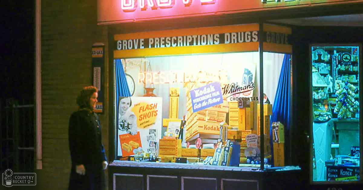 An adult female stands outside a 'Grove Prescriptions Drugs' store at night, wearing a dark coat with a high collar in an urban setting.