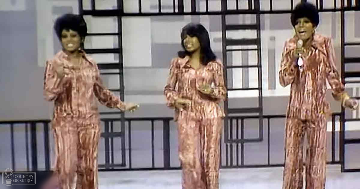 Three adult females in matching patterned jumpsuits perform on a brightly lit stage, likely from a music or variety show.