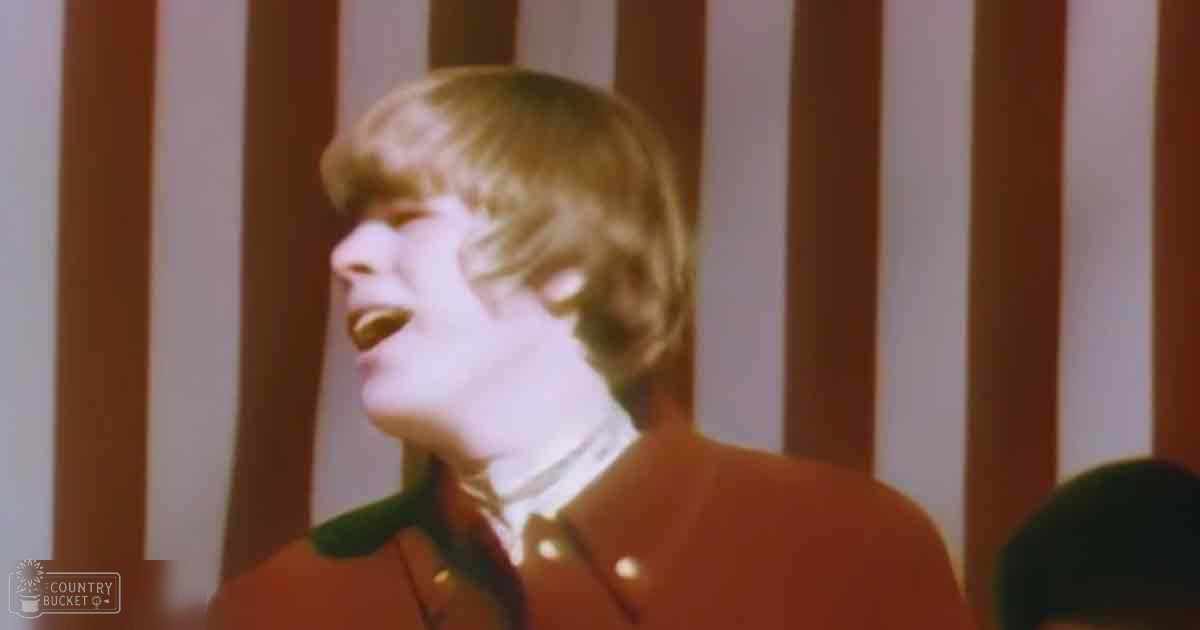 Young adult male singing indoors, wearing a red jacket with green accents and white shirt, natural lighting from behind.
