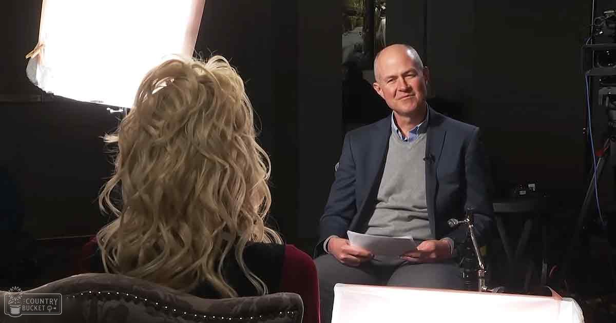 An adult female with long, wavy blonde hair sits in a red armchair during an interview setting; a middle-aged male wearing a gray sweater vest over a collared shirt and dark blazer holds papers seated on a white chair.