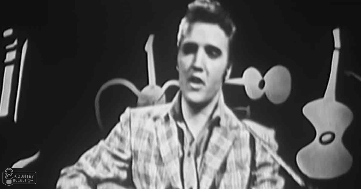 A young adult male singer in a mid-20th-century plaid shirt with rolled-up sleeves, standing on stage surrounded by musical equipment and instruments.