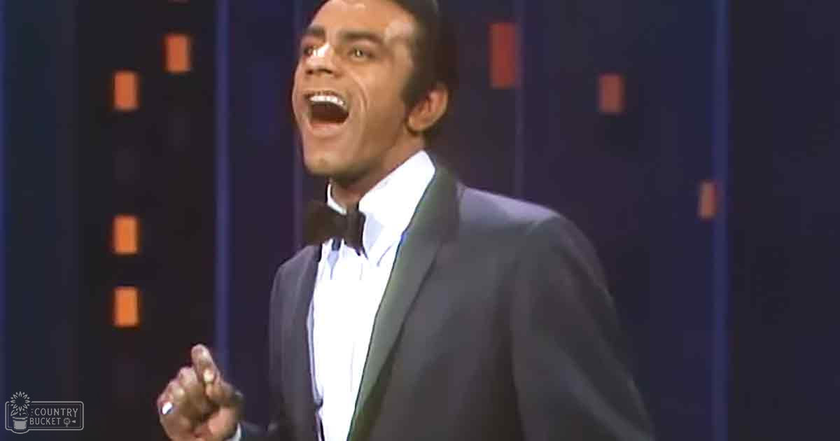 Adult male performer in a mid-20th-century dark suit jacket with white shirt and black bow tie, standing against an orange-squared backdrop suggesting a stage or studio setting.
