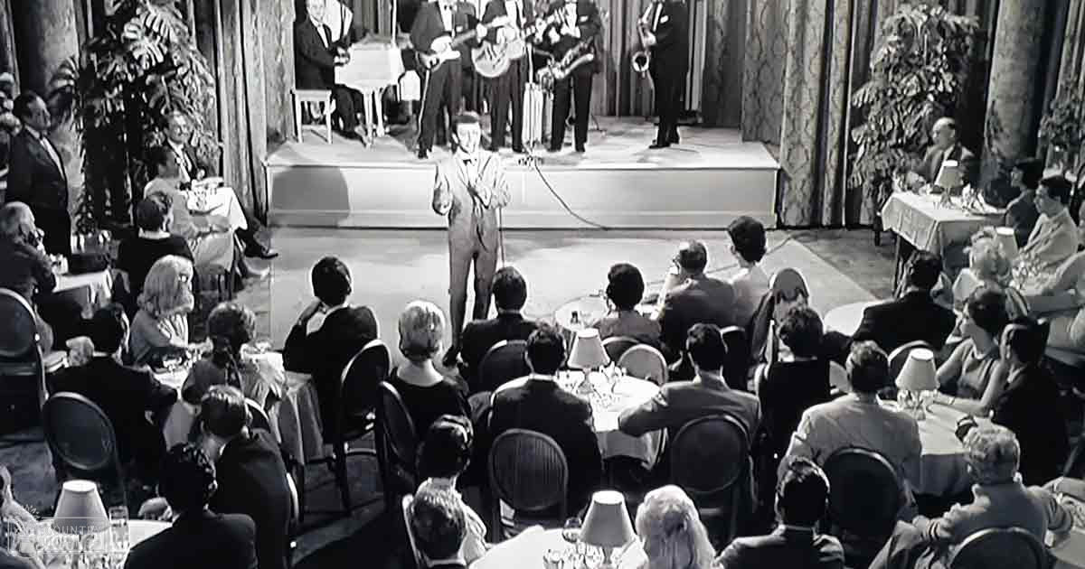 An adult male singer in a suit performing on stage amidst an audience dressed in formal attire at what seems to be a formal event or concert.