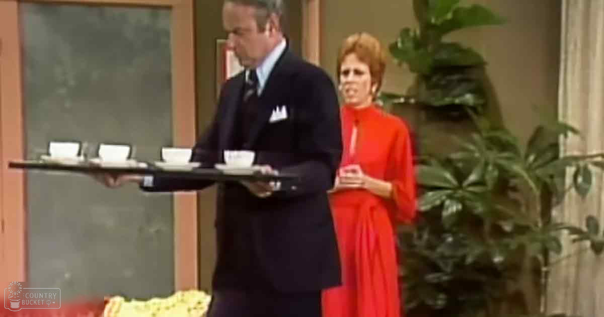 An indoor setting with a dark suit-clad male figure holding a tray of cups in front of a large green plant, with another individual wearing a bright red dress seated nearby.
