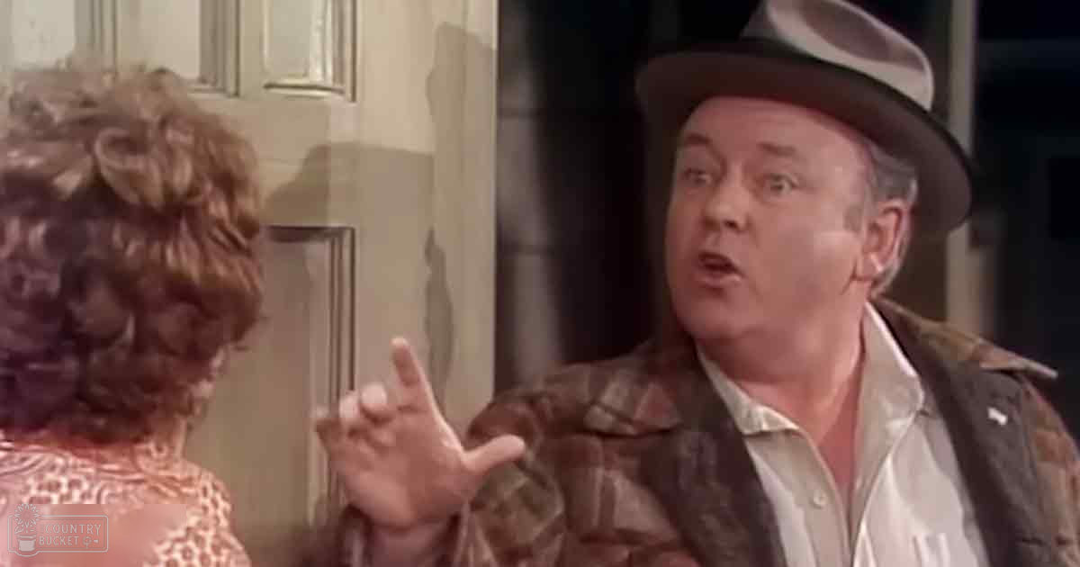 A middle-aged man wearing a brown plaid jacket and wide-brimmed hat indoors, with natural light creating shadows across the scene.