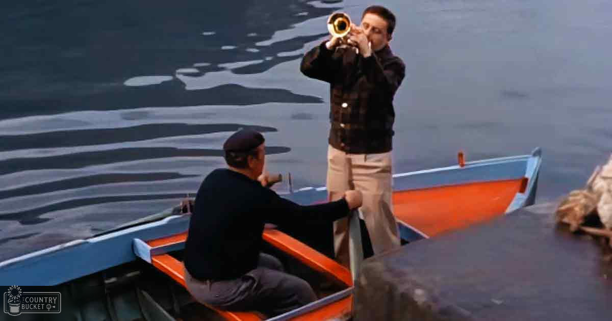 A young adult male plays a trumpet in a small rowboat on calm water; an older man sits nearby wearing a beret-style hat. A medium-sized dog with light-colored fur and darker patches is present.