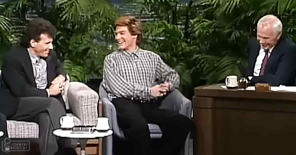 Talk show set with lush green foliage backdrop, two armchairs flanking a small round table holding a mug or cup and papers, suggesting an indoor studio environment. The setting is likely from the late 20th century.