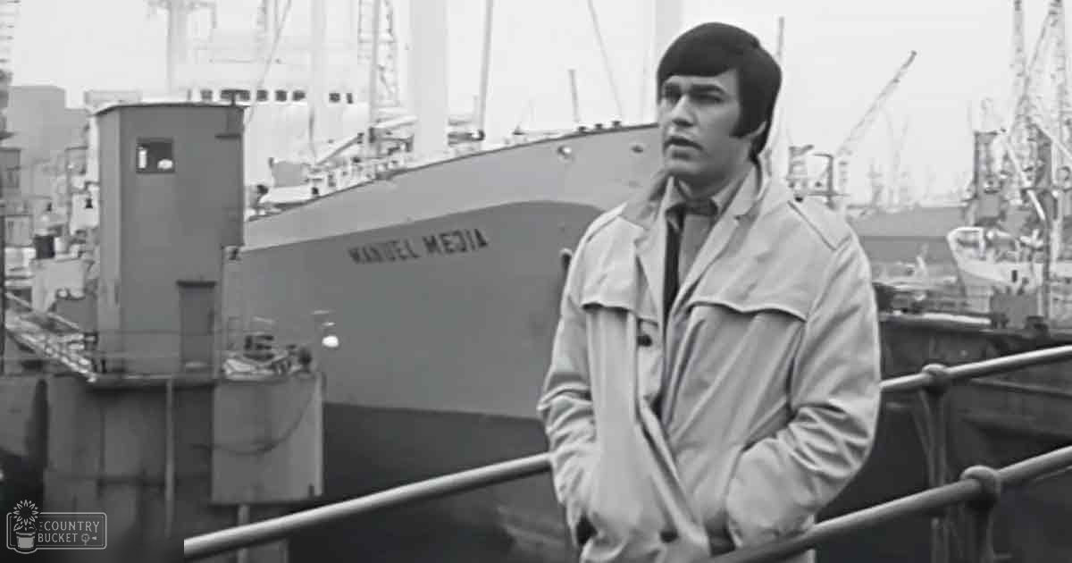 Young adult male in a beige trench coat at an industrial port with large ships and cranes, likely mid-20th century setting.