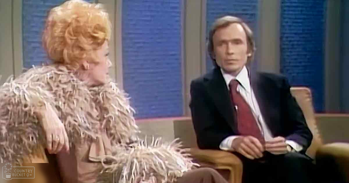 A woman with blonde updo and feathered garment, seated next to a formally dressed man on a light-colored armchair or couch in an indoor studio setting typical of the 1970s-early 1980s.