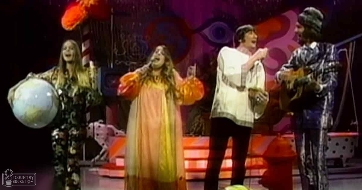 A performance scene with four adult female and male performers on a brightly lit indoor stage, wearing patterned and solid-colored outfits, holding a large globe in one corner.