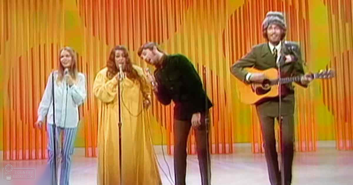 A musical performance with four adult performers dressed in vintage attire against a vibrant orange and yellow striped curtain.