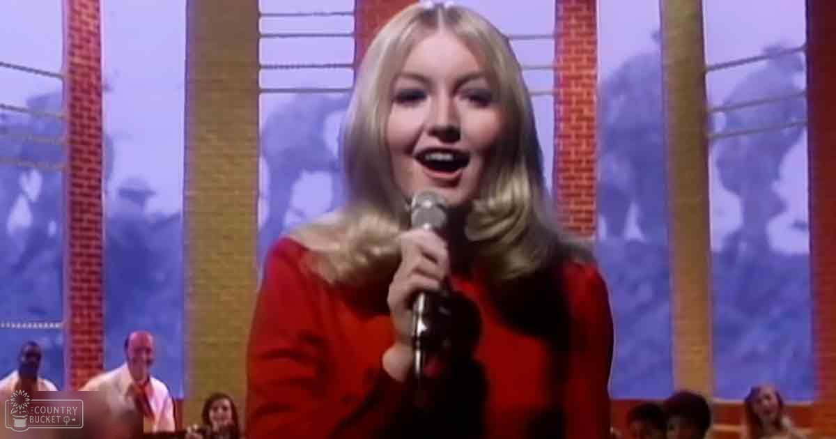 A young adult female singing into a microphone, wearing a red off-the-shoulder top in a studio setting with other adults in the background.