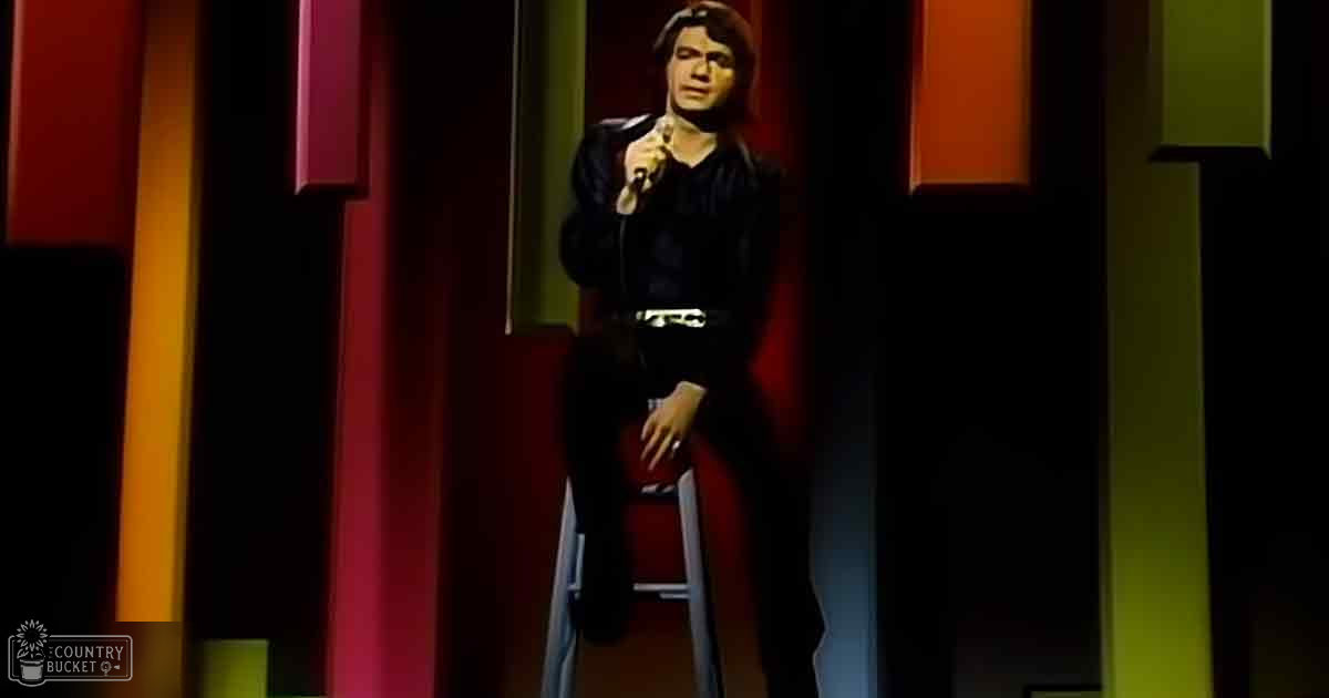Adult male performer in dark shirt and pants, seated on stool with microphone, against a red and yellow panelled background in an indoor studio setting.