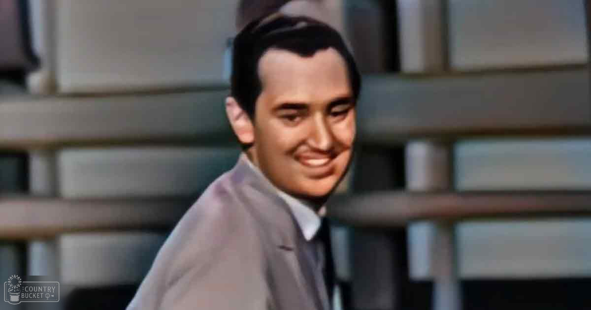 A young adult male wearing a light gray suit jacket in an urban setting with large windows and steps behind him, under natural daylight.
