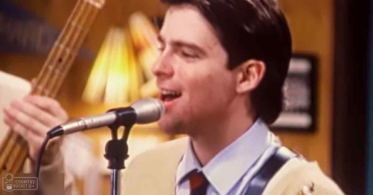 A young adult male in a light-colored suit jacket with a white shirt and red tie, holding a bass guitar on stage under warm lighting in an indoor performance space.