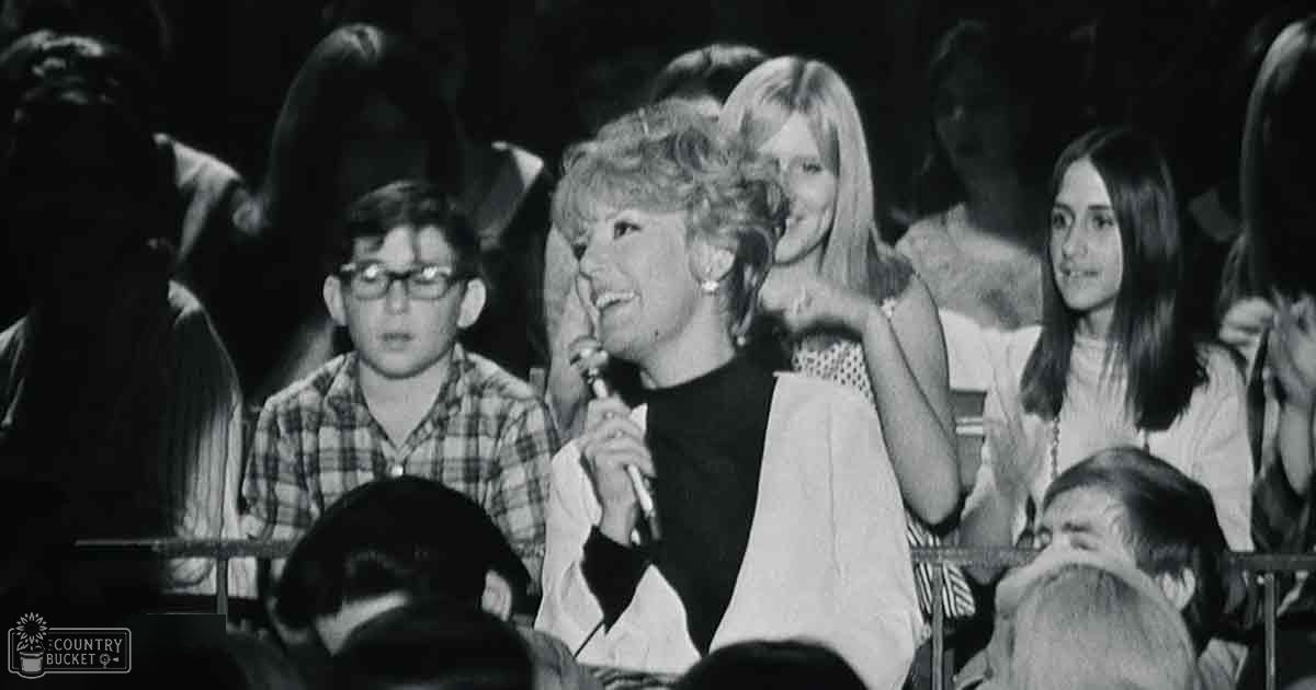 Adult female holding a microphone at an indoor event with dim lighting focused on the stage area.