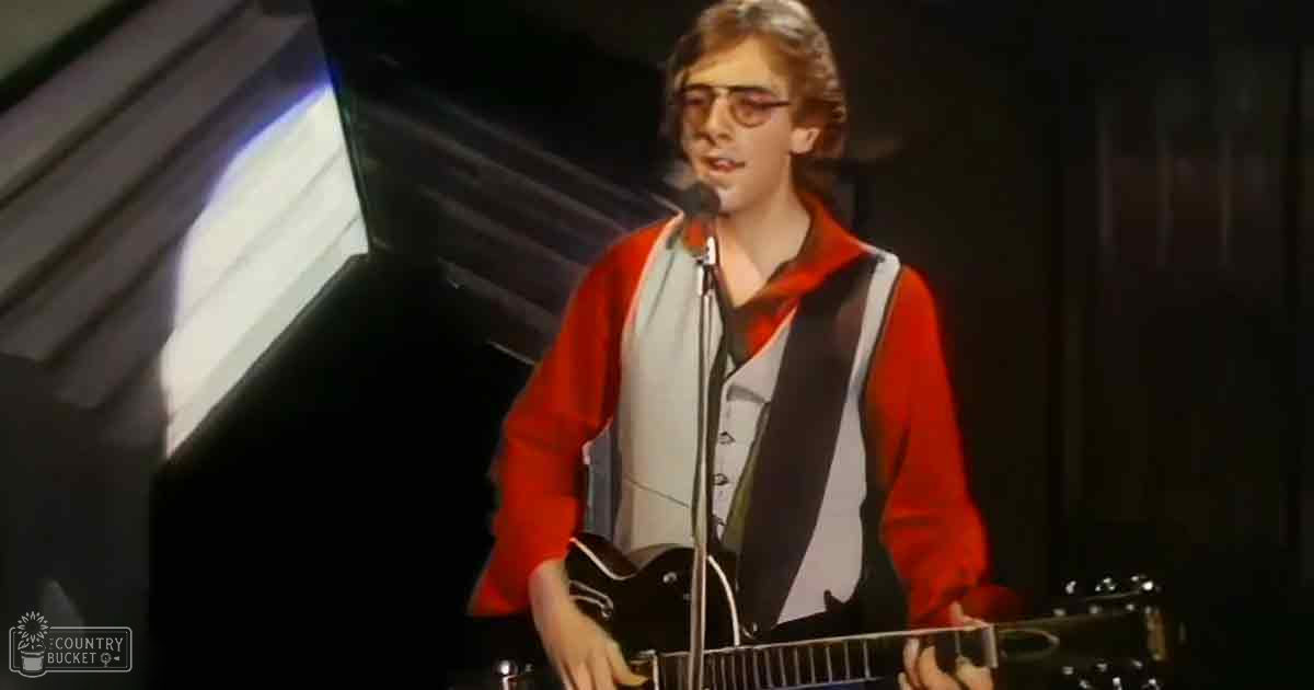 Young adult male in a red long-sleeved shirt with white accents and glasses performing on stage at an indoor venue.