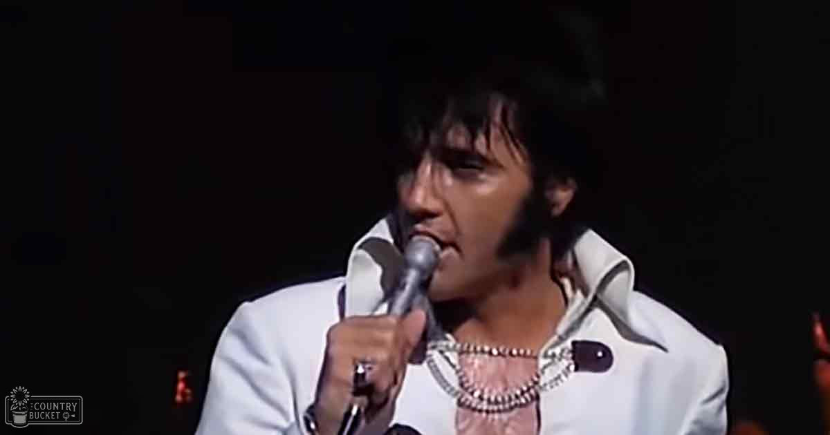 An adult male performer wearing a white suit with wide lapels and high collar, holding a microphone during what appears to be a concert or event at an indoor venue with red stage lighting.
