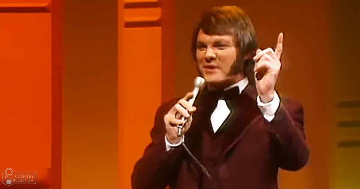 Adult male performer in a dark red suit with bow tie on stage, likely at a comedy club or theater during an indoor performance.