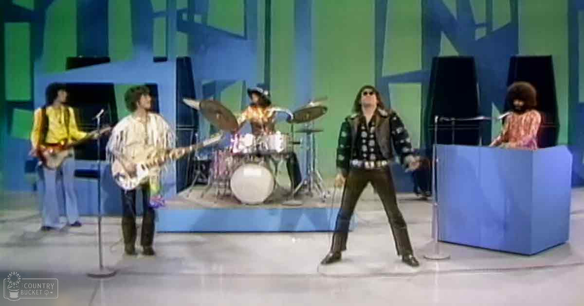 A musical performance scene in an indoor studio with musicians wearing patterned shirts and dark pants, a blue podium on the right, and a colorful geometric set background under even lighting.