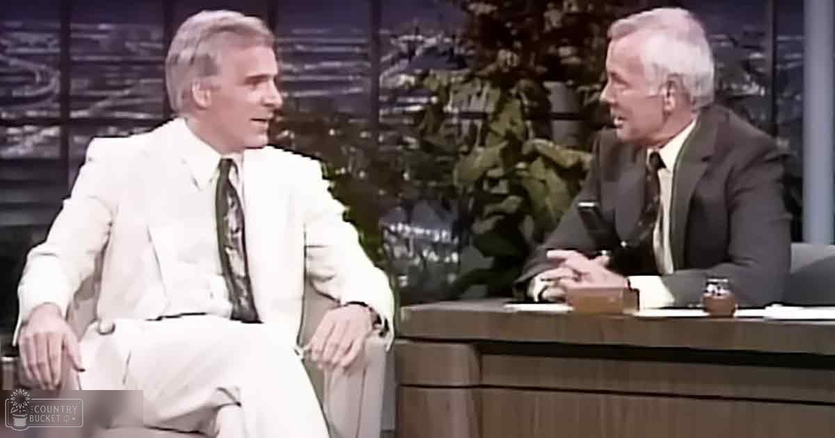 Two middle-aged men in a studio setting, one wearing a white suit with a patterned tie, the other in a dark suit and light shirt. A desk and cityscape background suggest an indoor talk show environment.