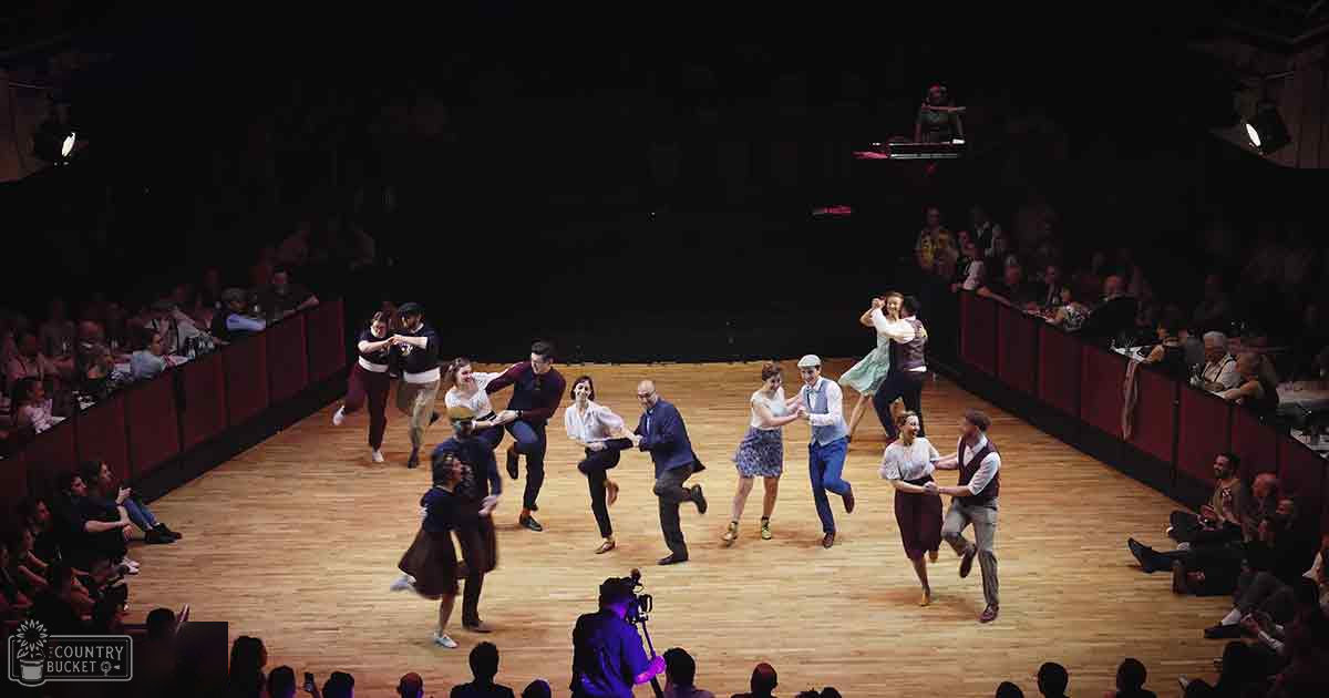 Young adults and adults performing a dance routine on a wooden floor with red barriers, audience seated around the stage, DJ booth at the back right corner under artificial evening lighting.