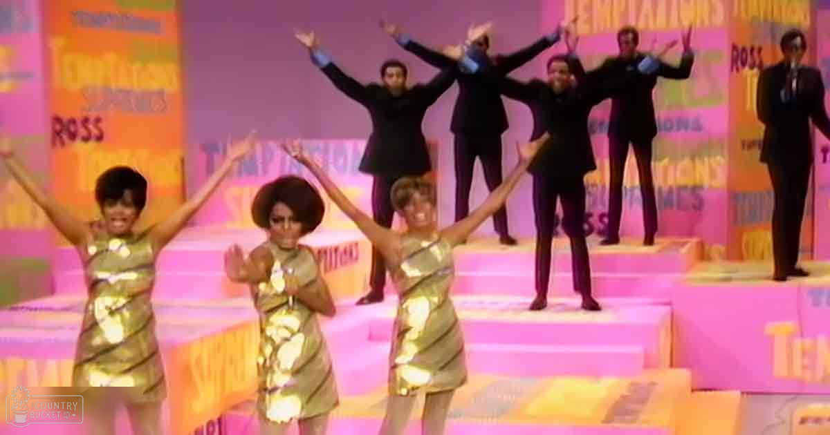 A performance scene with young adult females in gold striped dresses and synchronized dance moves on a stage with colorful backdrops with text.