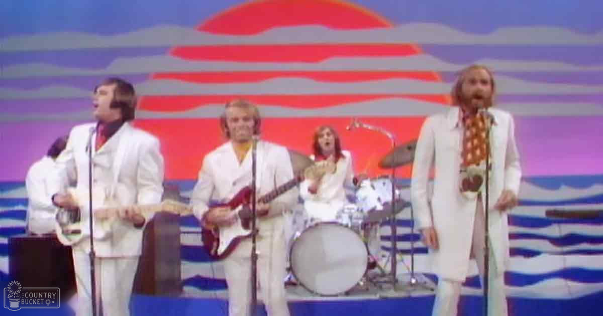 A 1970s musical performance on a stylized oceanic-themed stage with microphones and instruments, set against a backdrop of a stylized sunset.