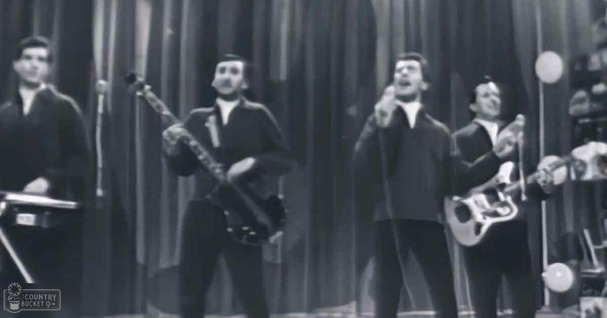 Musical performance scene with adult male singers and guitarists in front of a dark curtain under even lighting.