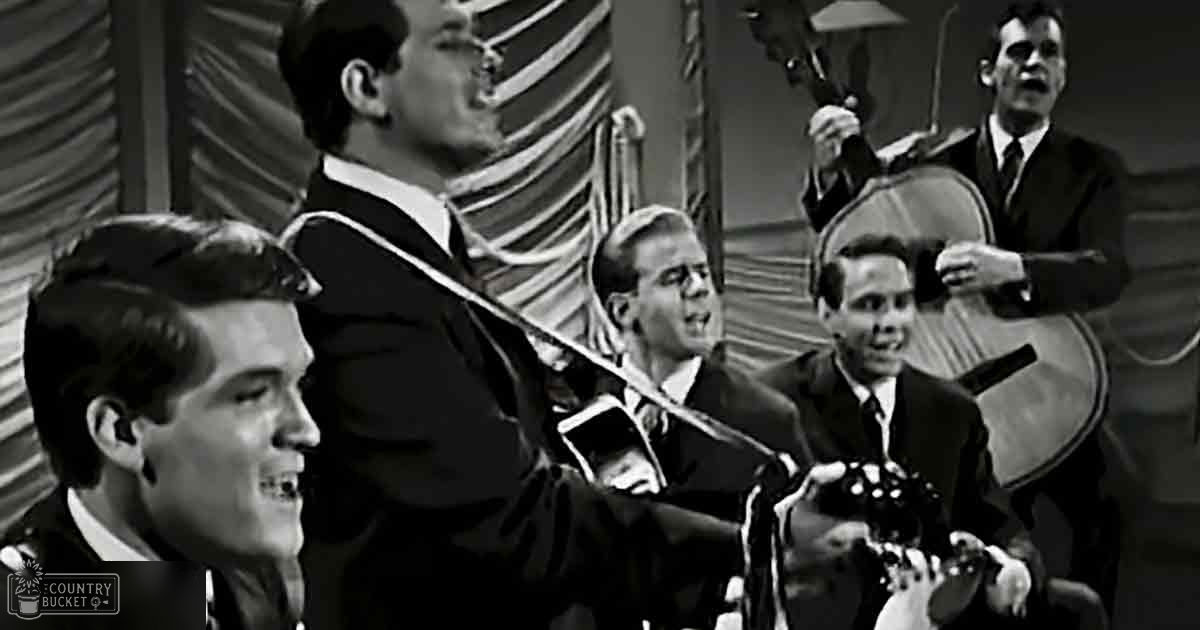 Musical performance scene in a studio or performance space with young adult males in dark suits, natural light from outside, vertical blinds and curtains, daytime setting.